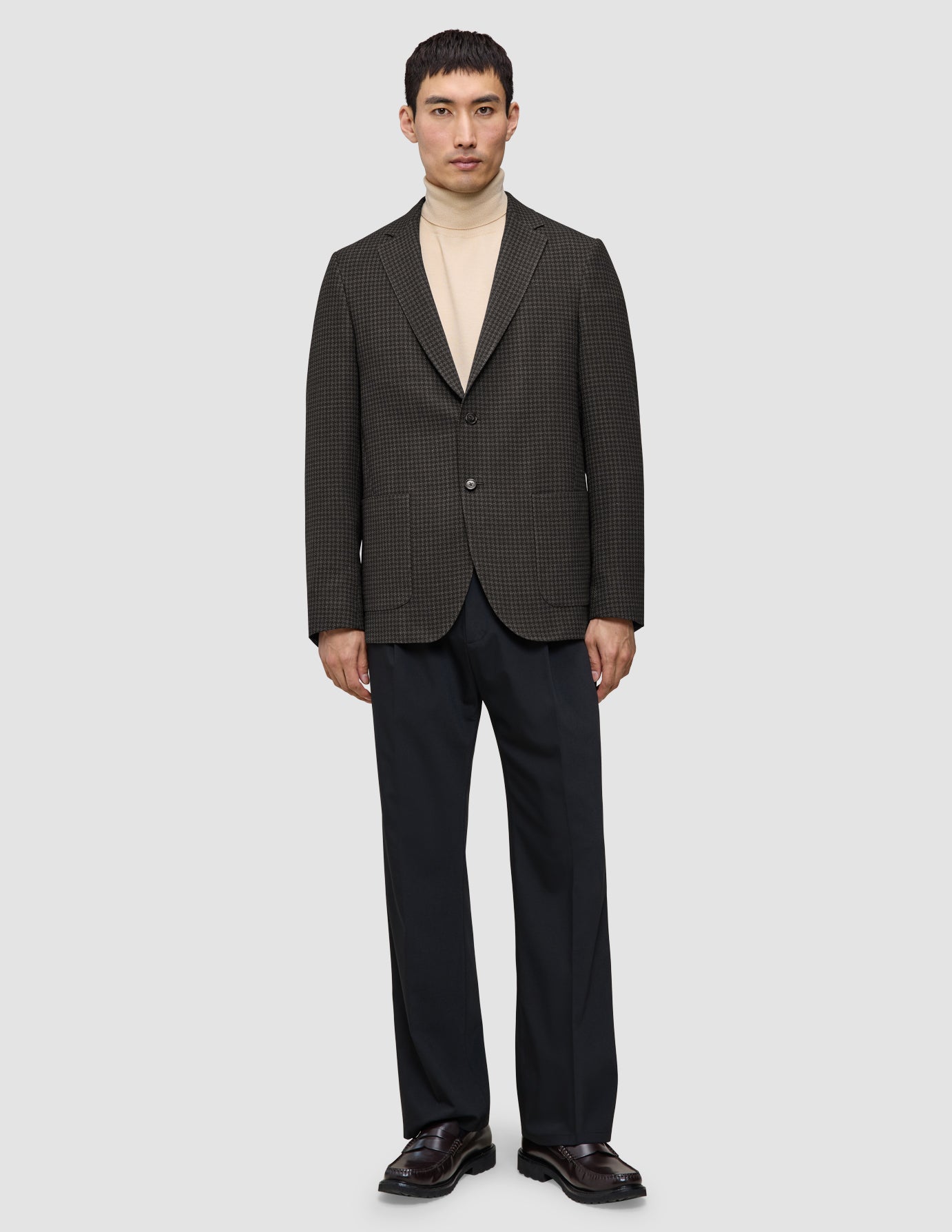 En person iført en mørkebrun, slim-fit Timeless Wool Blazer med tern over en beige rullekravebluse og mørke bukser.